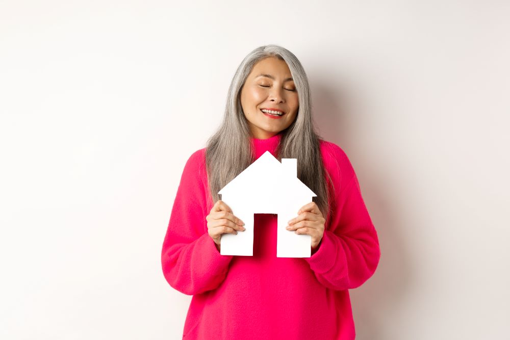 real estate beautiful dreamy asian lady hugging paper house model with closed eyes smiling as dreaming about buying apartment standing white background